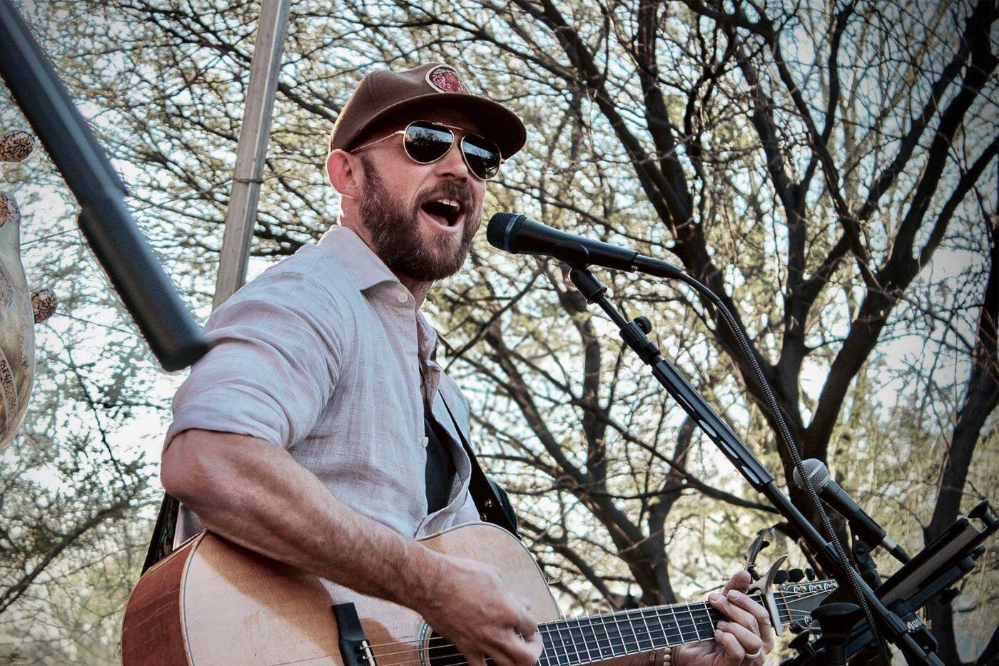Brett Dooley singing and playing acoustic guitar outdoors under bare desert trees
