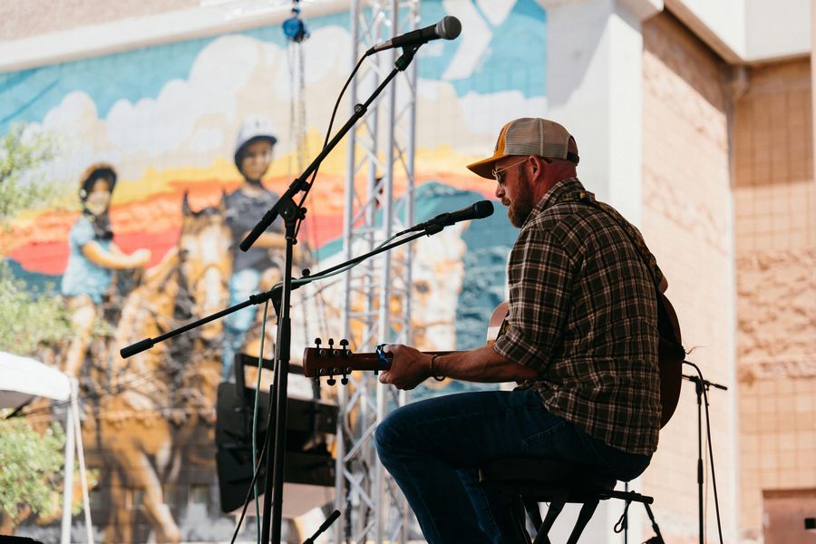 Brett Dooley playing acoustic guitar on an outdoor stage in front of a colorful mural
