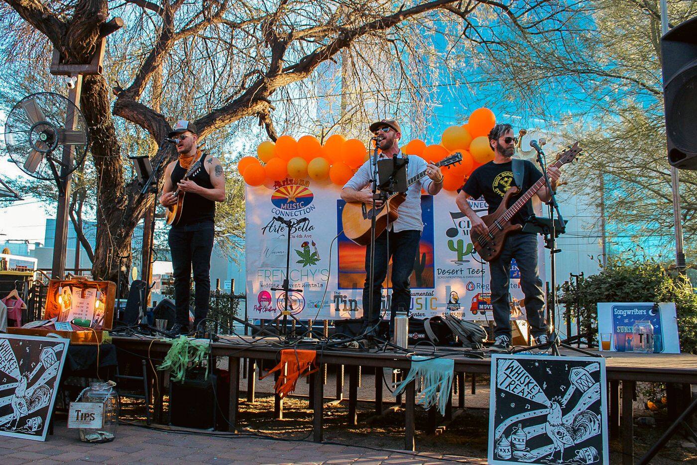 The Whiskey Treats playing mandolin, acoustic guitar, and bass on an outdoor stage, with rooster posters propped in front